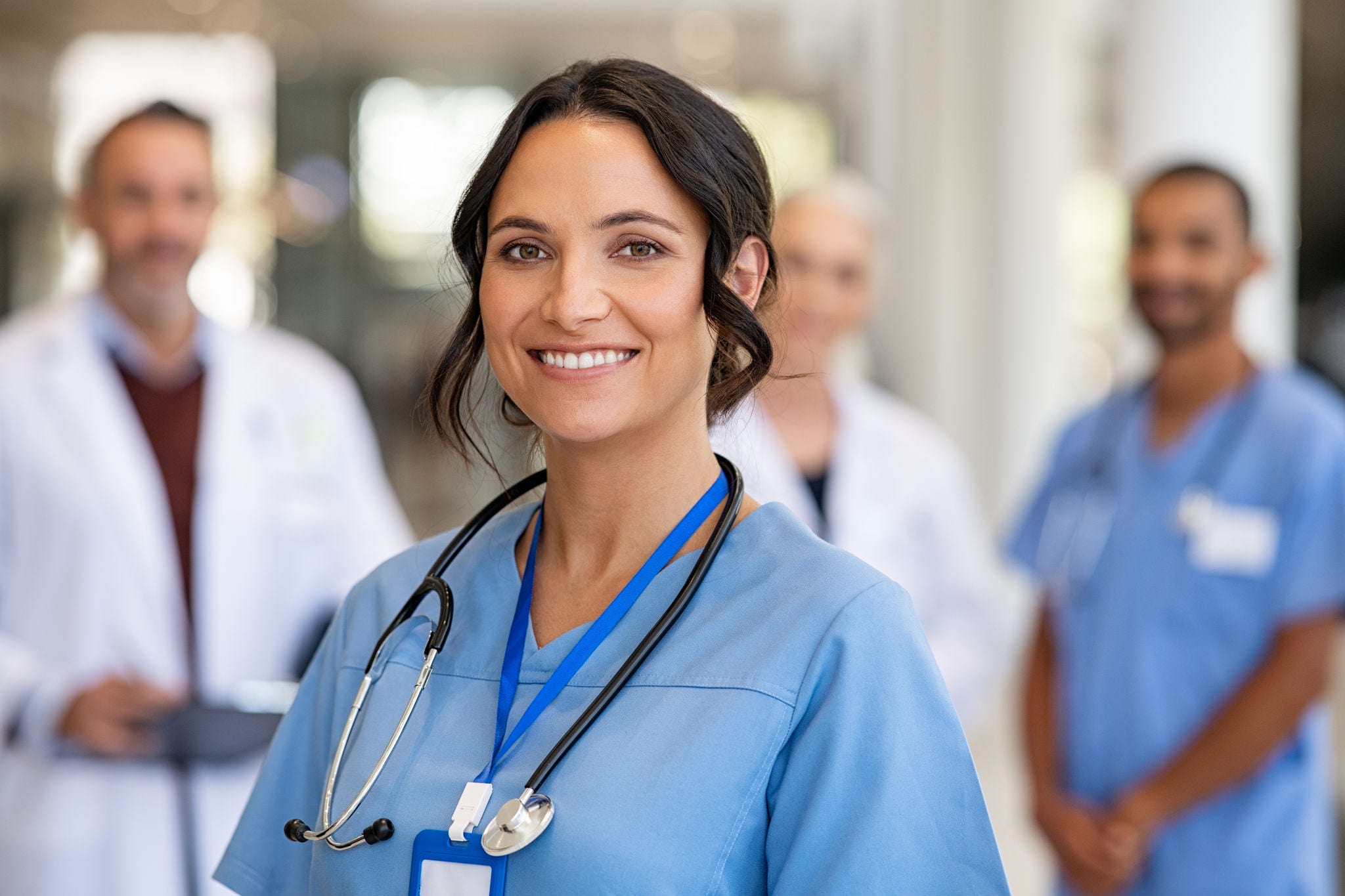 Portrait of happy young nurse in uniform with healthcare team in background. Successful team of doctor and nurses smiling. Beautiful and satisfied healthcare worker in private clinic looking at camera; Shutterstock ID 2054430419; purchase_order: -; job: -; client: -; other: -
