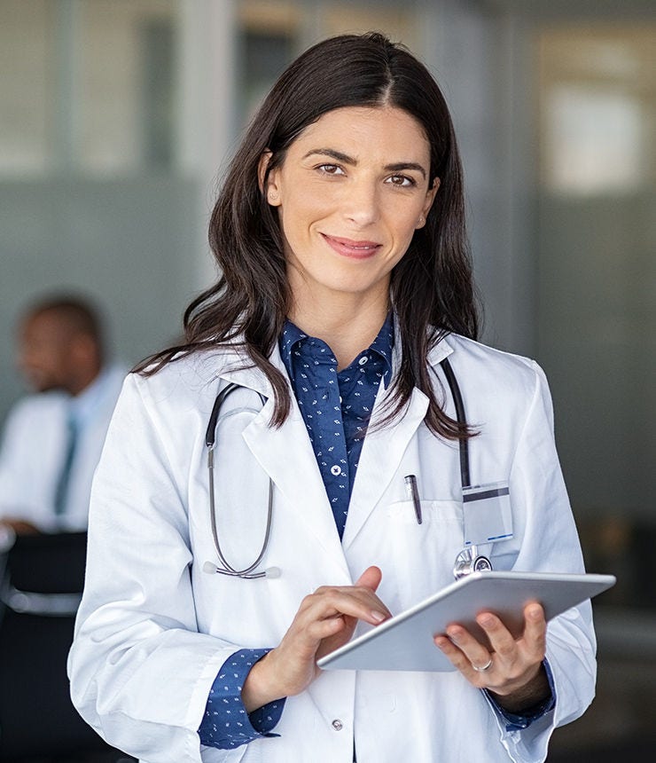 Portrait of beautiful mature woman doctor holding digital tablet and looking at camera. Confident female doctor using digital tablet with colleague talking in background at hospital. Latin nurse.; Shutterstock ID 1680655153; purchase_order: -; job: -; client: -; other: -