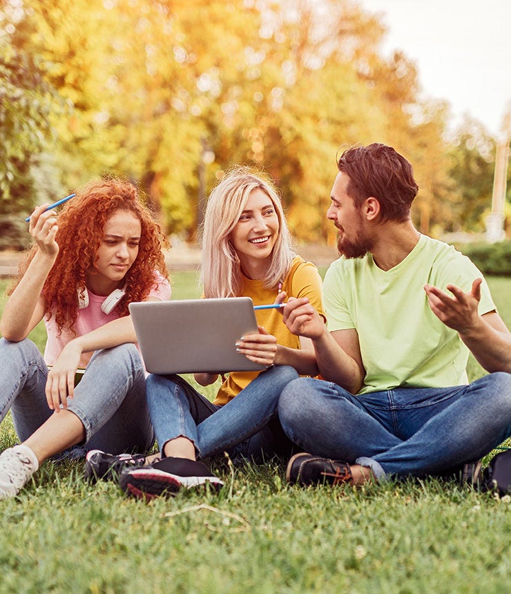 Young ginger woman reading assignment on laptop and thinking near cheerful classmates discussing project while doing homework on lawn in park together; Shutterstock ID 1587720022; purchase_order: -; job: -; client: -; other: -