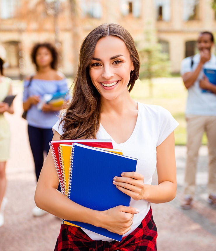 Focused shot of cheerful smart brunette caucasian girl is holding note books, smiling, standing near college building, her friends are behind, they passed tests, so cheerful and carefree!; Shutterstock ID 713217880; purchase_order: -; job: -; client: -; other: -