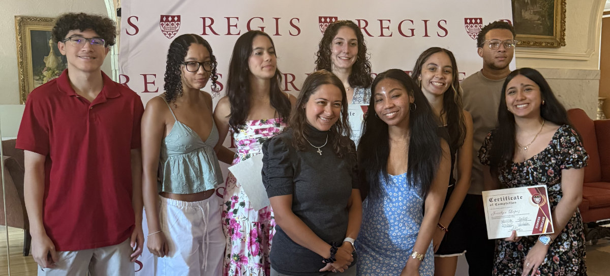 The 2025 cohort of Summer Scholars pose in front of a Regis backdrop