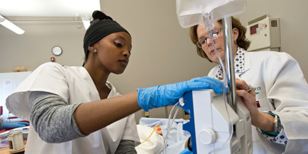 A Regis professor teaches a nursing student how to use an IV machine