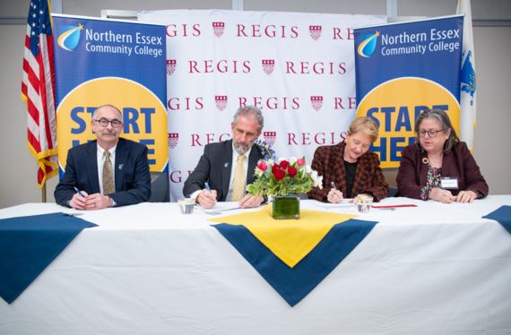Four administrators sit at a table signing the agreement in front of a Regis College back drop and NECC banner