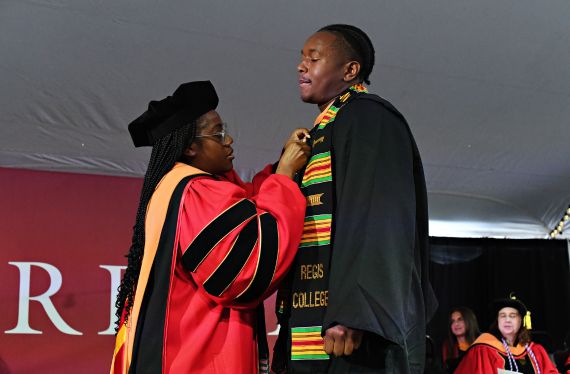 A faculty member in doctoral robes placing a pin on the lapel of a student in academic regalia