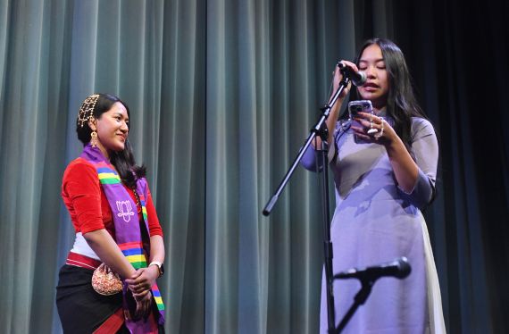 A student speaking into a microphone reading from a phone while another student looks on wearing an AAPI stole